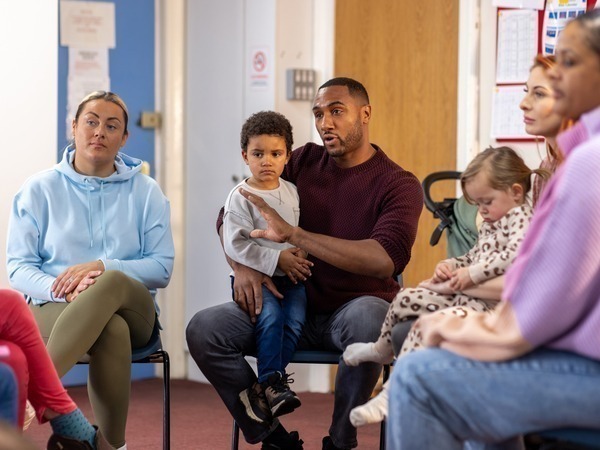 Over-the-shoulder shot of a father sitting down with his son on his lap. They are having a meeting with a childcare support worker out of shot and other parents in a community centre. 